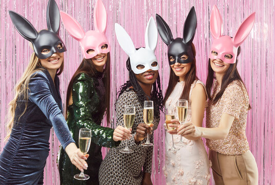 Group of joyful young women wearing bunny masks posing with glasses of champagne. Happy female friends celebrating special occasion during carnival or bachelorette party against shiny pink background. - Powered by Adobe