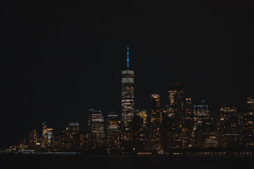 The illuminated Lower Manhattan skyline and One World Trade Center glittering at night, seen from the water.