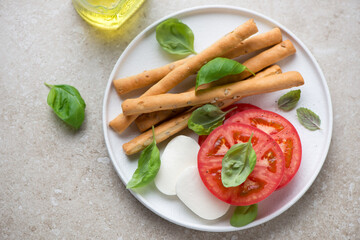 Salted breadsticks and ingredients for caprese on a white plate, horizontal shot on a beige stone background, high angle view