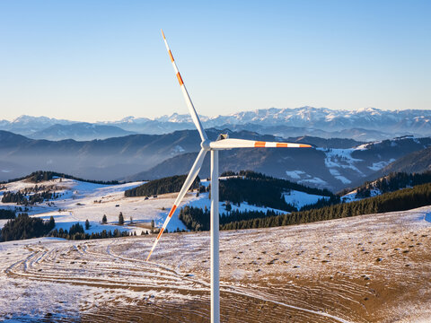 Aerial view of a towering wind turbine stands stark against the snowy hills and distant, hazy mountains, Sommeralm, Steiermark, Austria.