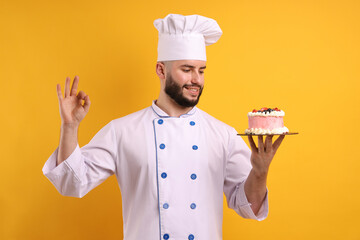 Happy confectioner in uniform holding cake with berries and showing ok gesture on orange background