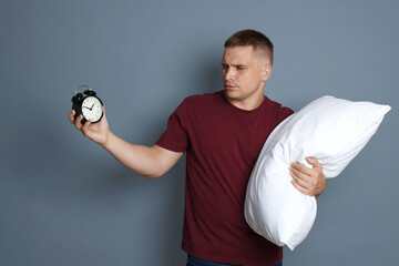 Man with alarm clock and pillow suffering from hangover on grey background