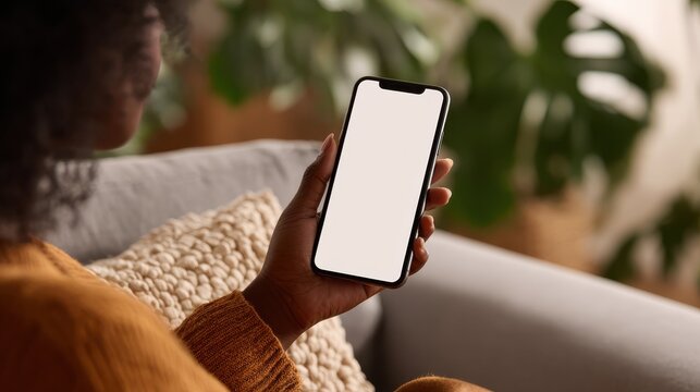 young black african woman holding smartphone in hand with white black empty mockup screen template using cell phone relaxing on sofa at home over shoulder view mobile apps advertising concept no logo