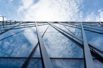 Tall modern building with glass facade and metal beams reflecting clouds and blue sky, viewed from...