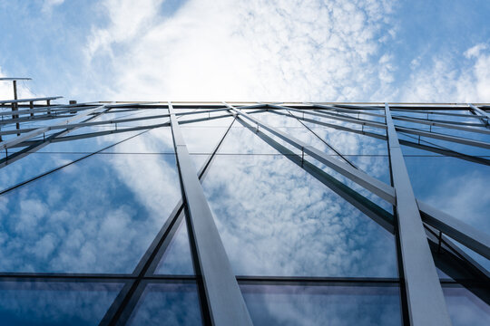 Tall modern building with glass facade and metal beams reflecting clouds and blue sky, viewed from below. Suitable for illustrating architecture, urban development, or corporate design concepts.
