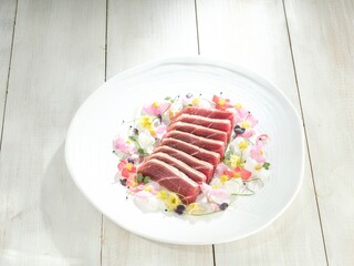 Slices of raw tuna rest beautifully on a white plate, surrounded by a delicate arrangement of pink, yellow, and white petals and greens. It looks artful and delicious