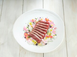 A close-up shows fresh, sliced ahi tuna arranged with edible flowers and greens on a pristine white dish, set against a light-colored wood tabletop for an elegant presentation