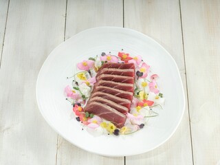 Seared tuna steaks, sliced beautifully, rest upon a scattering of colorful edible flowers. This elegant dish is presented on a white ceramic plate atop a white wooden surface