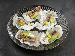 Four portions of tuna, avocado and lemon are presented on delicate rice paper on a textured plate. Black sesame seeds and tiny white flowers add to the appeal