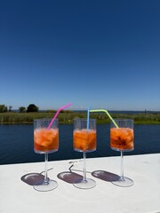 Three orange summer cocktails with ice and colorful straws sit on a white ledge against a clear blue sky and waterfront