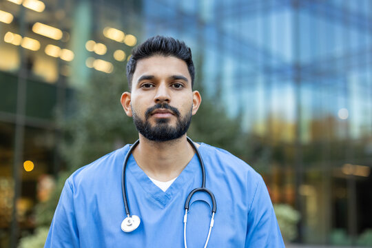 An Indian healthcare professional stands confidently in front of a blurred architectural backdrop, with a stethoscope around his neck.