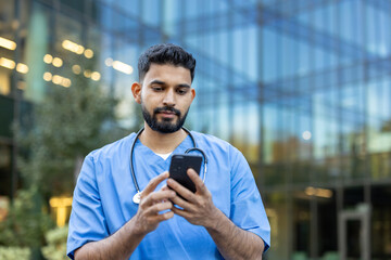 A male healthcare professional in scrubs using his smartphone outdoors, near a modern building.