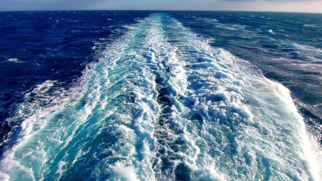 Wake. A view of ocean waves created by a passing cruise ship. The water is deep blue with white frothy wake trailing behind. Bright sunlight reflects on the surface.