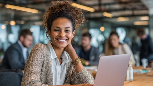 smiling young african american businesswoman working on a laptop at her desk in a bright modern office with colleagues in the background no logos no brands ar 169