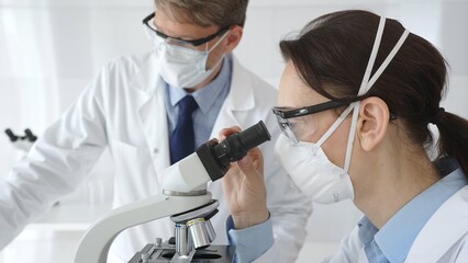 Two scientists in lab coats and safety gear conducting research, one observing through a microscope and the other holding a test tube with turquoise liquid. Medicine and science concept