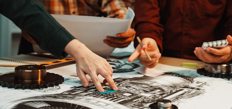 A focused team of engineers collaborates on intricate technical drawings of mechanical engine components, showcasing the importance of teamwork in a modern workshop environment. SACTR