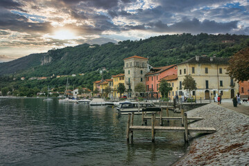Pella, Italy: The picturesque waterfront of the village on Lake Orta, with colorful historic buildings and small boats moored against a backdrop of forested hills under a dramatic sky.