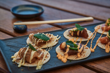 A Platter of Gourmet Snack Bites Featuring Fresh Fish and Unique Sauces Served on a Slate Board with Wooden Chopsticks and Saucer