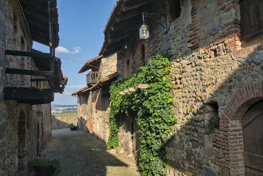 Ricetto di Candelo, Italia: Una stretta strada di ciottoli nel borgo medievale con muri in mattoni e pietra coperti di edera, illuminata dal sole sotto un cielo azzurro.