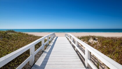 Obraz premium white wooden boardwalk leading to a tranquil beach under a clear blue sky
