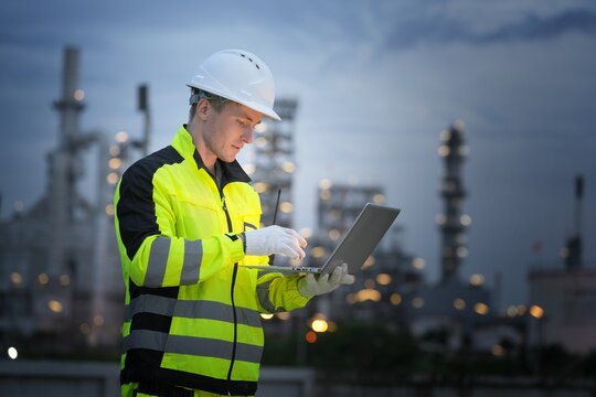 Worker in safety gear using laptop at industrial site during twilight - Powered by Adobe