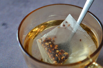 Close-up of a transparent cup with herbal tea and a tea bag steeping inside. Natural lighting and visible tea particles emphasize freshness and simplicity.