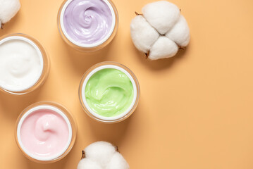 Colored organic body creams in jars on yellow pastel background decorated with cotton flowers, top view