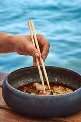 A Hand Gently Using Chopsticks to Enjoy a Delicate Dish by the Water, Showcasing the Art of Dining and Culinary Experience in a Serene Setting