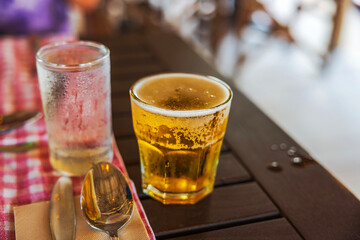 Close up view of cold beer with foam and condensation in glass on wooden table with spoon and napkin. Willemstad. Curacao.