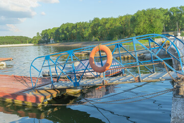 Rustic blue painted metal pier with lifebuoy tied on railing leading to peaceful river with moored covered motor boat and dense trees under clear sky. Blue metal pier with life buoy near river.