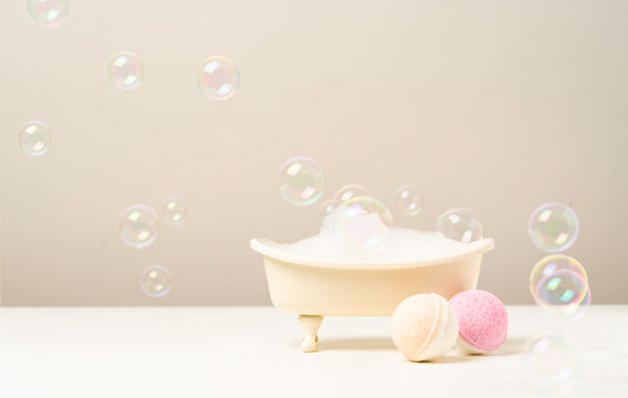 Miniature bathtub filled with foam, surrounded by bath bombs and floating soap bubbles. Concept of spa, self-care and relaxation in soft neutral tones. - Powered by Adobe