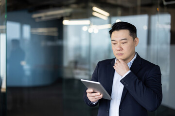 A thoughtful young Asian man is standing inside an office and looking seriously at the screen of a tablet he is holding in his hand