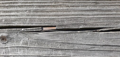 A close-up of weathered wood with faded gray tones, fine grain lines, and a deep horizontal crack. The rough, aged surface creates a rustic, organic wooden texture background