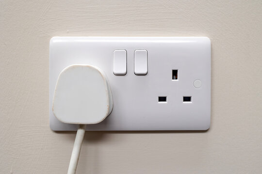 White UK 3-pin mains plug inserted into modern dual wall socket with switches. Mounted on a light painted wall. Suitable for illustrating household electricity, safety, and electrical infrastructure