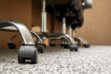 Low-angle close-up of caster wheels and chrome bases of office chairs lined up beside a boardroom table on patterned carpet flooring, with defocused background. For business and workplace themes