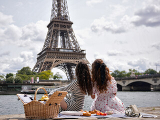 Two women sit on picnic blanket facing Eiffel Tower across the river. 