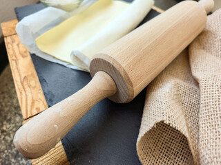Raw puff pastry on a black board, close-up. Nearby is a wooden rolling pin and flour. Concept of home baking or dough preparation.