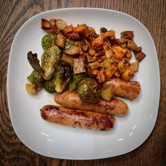 Dinner plate with grilled sausages, roasted Brussels sprouts, and a colorful hash of diced vegetables including sweet potatoes, onions, and bacon on a wooden table