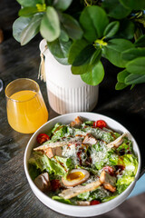 Fresh salad with grilled chicken, cherry tomatoes, grated cheese, and soft-boiled egg in white bowl, served with orange juice on rustic wooden table.