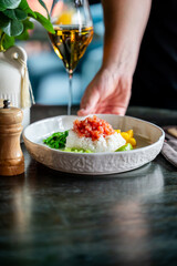 Hand serving a bowl of rice with tomatoes, vegetables, and lime on a table with white wine and seasoning, creating a fresh and elegant dining atmosphere.