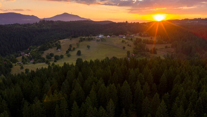Drone image captures the last sunlight spilling across a dense conifer forest and meadows at the edge of sunset