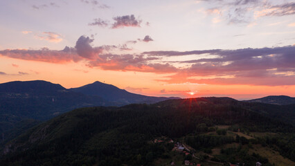 Sun sets behind distant ridges, painting the clouds with soft orange, pink, and purple tones above dark green forests