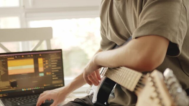 Musician composing music at home, working on laptop with audio editing software and holding blue electric guitar. Creative music production process in cozy daylight home studio, playing the guitar.