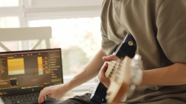Young musician composing music at home, working on laptop with audio editing software and holding blue electric guitar. Creative music production process in cozy daylight home studio
