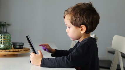Curious little boy touches screen of tablet with finger searching game. Concentrated kid carefully uses modern device sitting at desk at home