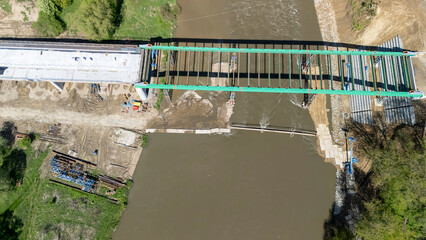 Aerial view of a bridge under construction over a river, showcasing workers and heavy machinery at a construction site with greenery in the background.