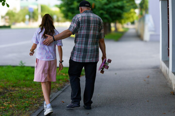 Obraz premium Grandfather walks with granddaughter down a calm street while she carries a skateboard