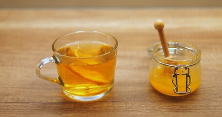 Glass mug of brewing tea with swirling slice of lemon near honey jar on wooden surface. Refreshing beverage infused with sweet and sour flavours