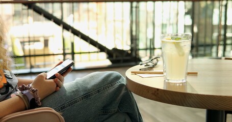 Young woman engages with modern mobile phone sitting at table on restaurant terrace. Fresh lemon beverage with straw and sunglasses on table side view