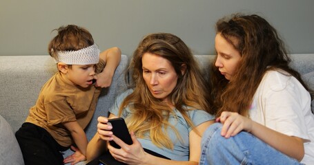 Blonde mother sits on couch with children looking at smartphone. Preschooler boy watches with curiosity and teen girl leans in talking to woman on sofa